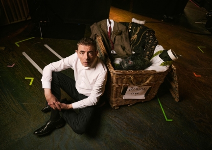 Rowan Atkinson sitting on a stage floor beside a wicker costume basket labelled Rowan Live, with costumes from his famous characters piled inside